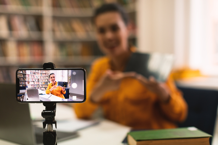 Mulher gravando um vídeo educativo em uma biblioteca, usando o storytelling para apresentar conteúdo enquanto aparece enquadrada pela câmera do smartphone.